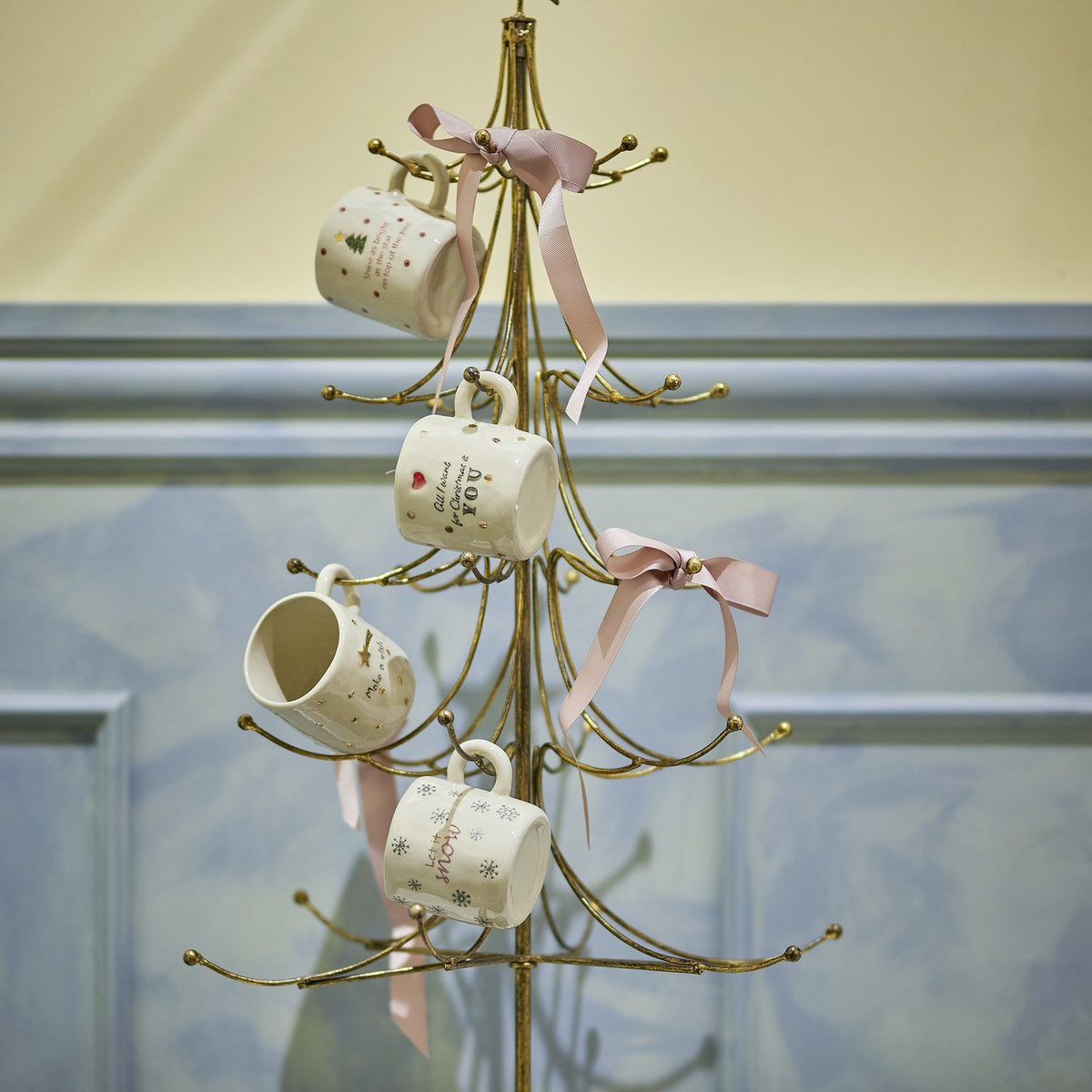 Wen Iron Christmas Decoration in gold, a tree-shaped metal stand with curved tiered arms and hook tips, displaying four mugs tied with pink ribbons.