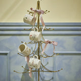 Wen Iron Christmas Decoration in gold, a tree-shaped metal stand with curved tiered arms and hook tips, displaying four mugs tied with pink ribbons.