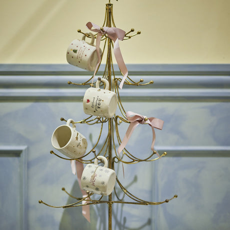 Wen Iron Christmas Decoration in gold, a tree-shaped metal stand with curved tiered arms and hook tips, displaying four mugs tied with pink ribbons.