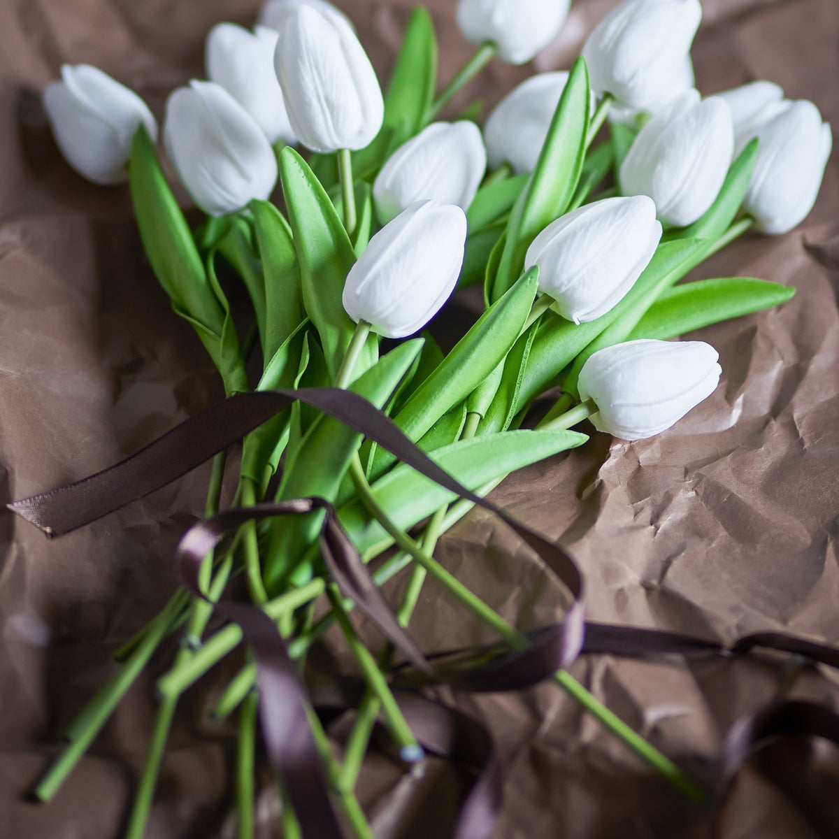 Artificial white tulip bouquet with closed petals and long green stems and leaves, tied with a brown ribbon on crumpled brown paper.