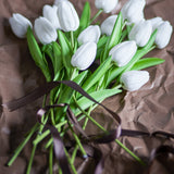 Artificial white tulip bouquet with closed petals and long green stems and leaves, tied with a brown ribbon on crumpled brown paper.