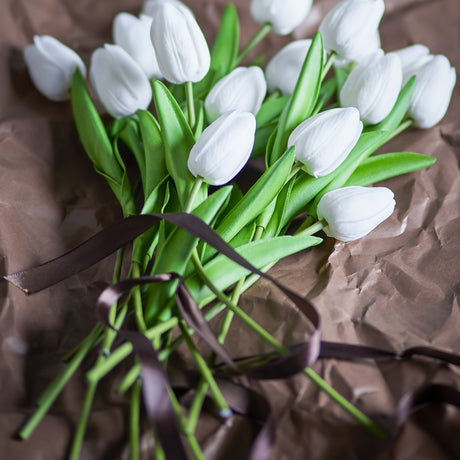 Artificial white tulip bouquet with closed petals and long green stems and leaves, tied with a brown ribbon on crumpled brown paper.