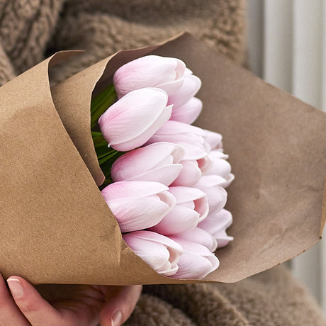 Tulip Artificial Bouquet in pink, closed cupped blooms wrapped in brown kraft paper, held by hand against a neutral indoor background.