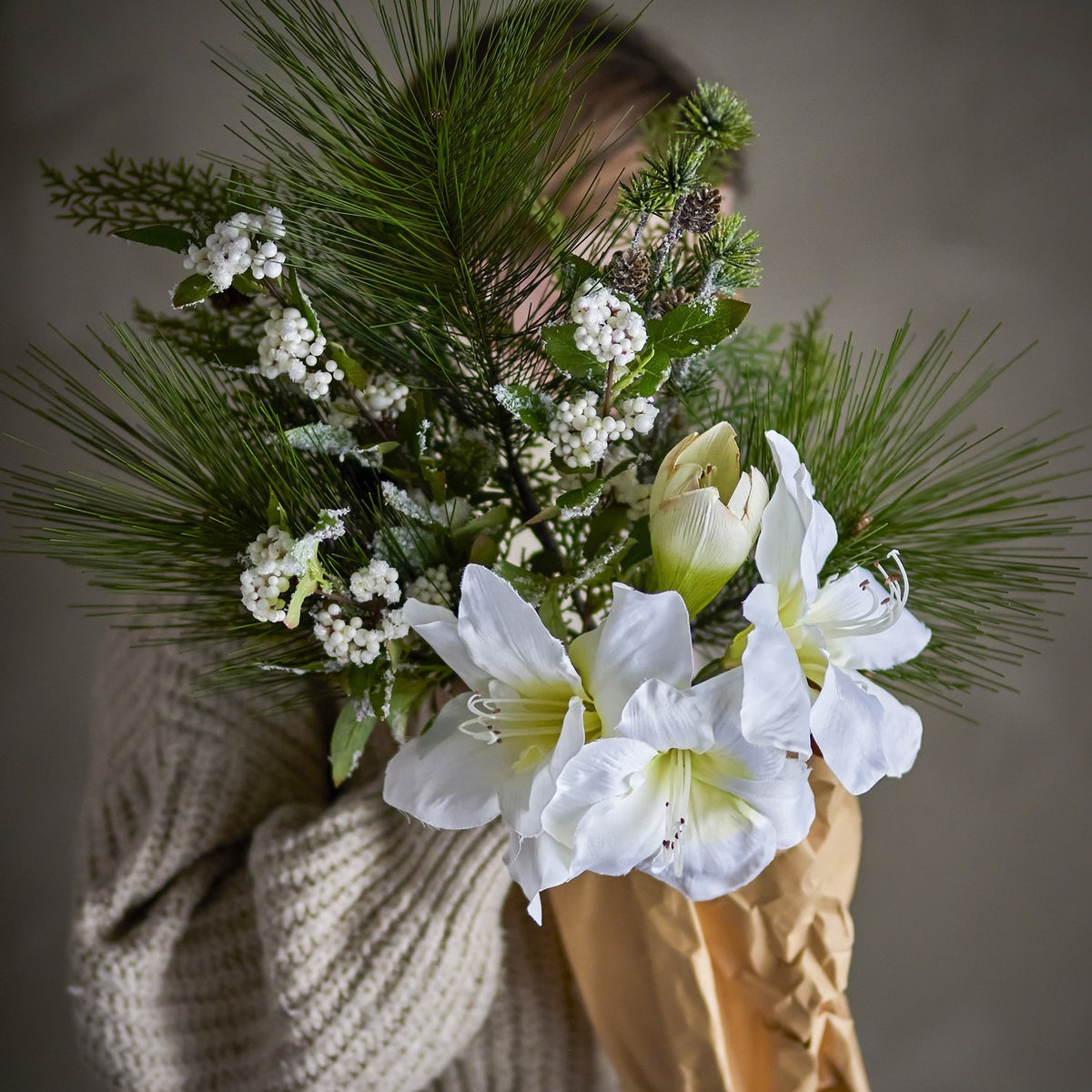 Kristin Artificial Christmas Bouquet in White, held against a sweater, showing white trumpet blooms, pine needles, frosted leaves and berry clusters wrapped in brown paper.