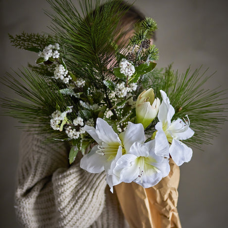 Kristin Artificial Christmas Bouquet in White, held against a sweater, showing white trumpet blooms, pine needles, frosted leaves and berry clusters wrapped in brown paper.