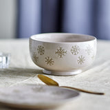 Beige stoneware bowl from the Snowie Christmas set, 13cm, with raised snowflake motifs around the exterior and a plain interior, shown on a table with a gold spoon and matching plate.