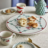 Yule Stoneware Christmas Lazy Susan in white, round ceramic turntable with fir garland and heart motifs, shown on a dining table with cookies, mugs and a green teapot.