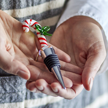 Lynn Christmas wine stopper held in hand, featuring a red and white striped glass candy cane handle with a green glass bow, set on a stainless steel cone with ridged silicone grip.