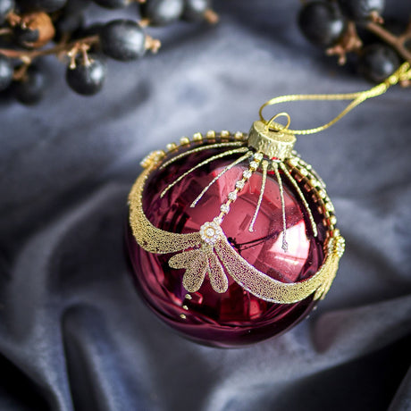 Round glass Christmas bauble from the Suman set in glossy red, featuring a glittering gold swag with beaded lines and leaf-shaped drops, shown against a dark fabric backdrop with berries.