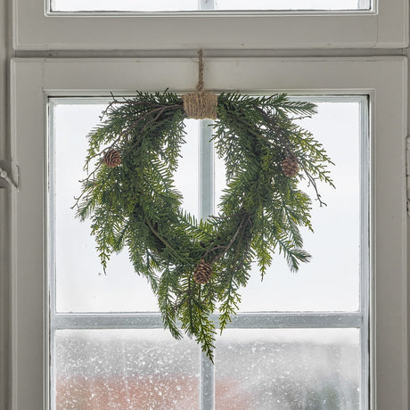 Pine Heart Christmas Decoration in green, a heart-shaped artificial pine wreath with small pinecones and a jute hanging loop, shown hanging in a window.