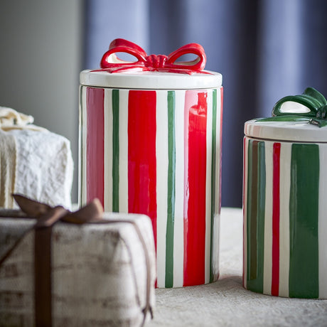Marnia Striped Christmas Storage Jar, a cylindrical stoneware canister with red, green and white vertical stripes and a glossy red bow on the lid, shown beside a smaller green-bow version.