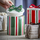 Marnia Christmas storage jar in green striped ceramic with lid lifted off, showing a hand-modelled green bow handle, styled beside a matching red striped jar and wrapped gifts.