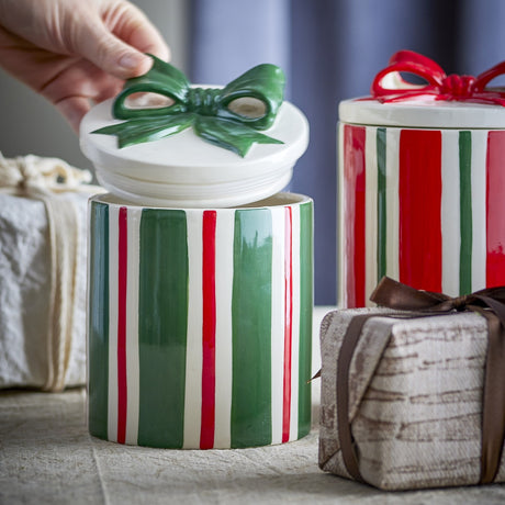 Marnia Christmas storage jar in green striped ceramic with lid lifted off, showing a hand-modelled green bow handle, styled beside a matching red striped jar and wrapped gifts.