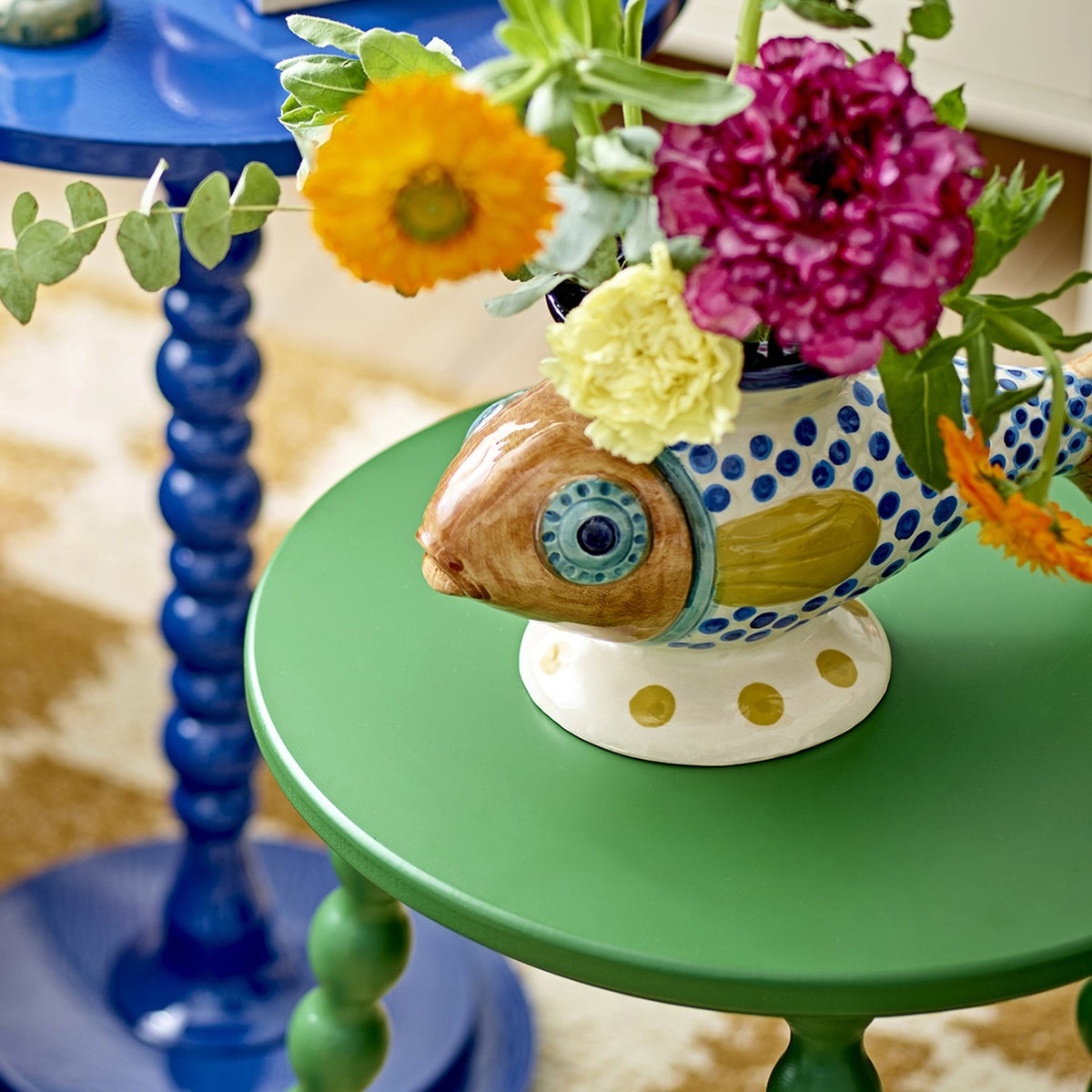 Nobu Fish Stoneware Vase in multicoloured hand-painted ceramic with blue dotted scales and fin, holding fresh flowers on a green side table beside a blue table.