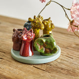 Piero Stoneware Vase 10.5cm in Multi, a round ceramic base with six animal-head openings in red, green, brown and amber glazes, shown on a wooden surface with pink blossom.