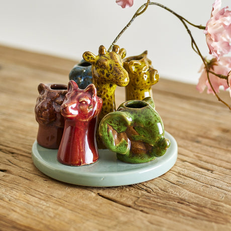 Piero Stoneware Vase 10.5cm in Multi, a round ceramic base with six animal-head openings in red, green, brown and amber glazes, shown on a wooden surface with pink blossom.