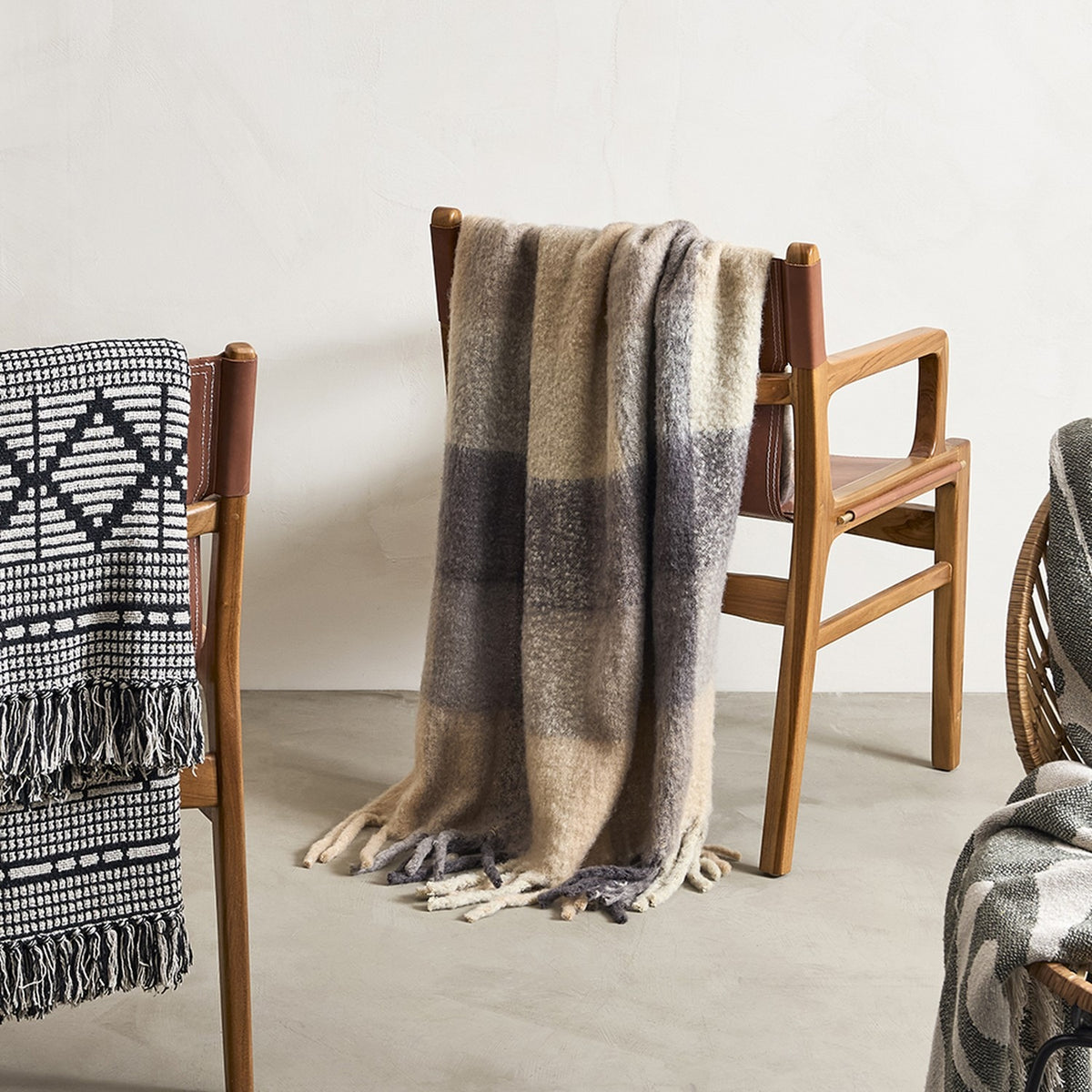 Hamisha Grey Throw in multicoloured block check with fuzzy texture and long fringe, draped over a wooden armchair with leather straps.