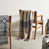 Hamisha Grey Throw in multicoloured block check with fuzzy texture and long fringe, draped over a wooden armchair with leather straps.