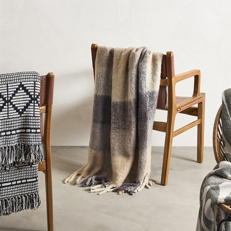 Hamisha Grey Throw in multicoloured block check with fuzzy texture and long fringe, draped over a wooden armchair with leather straps.