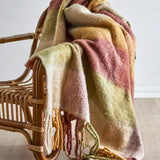 Hamisha Throw in Multi draped over a rattan armchair, showing a broad striped check in rust, ochre, sage, and cream with twisted fringe ends, against a plain background.