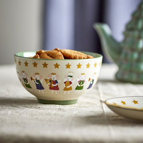 Cream stoneware Christmas bowl with green rim and base, decorated with angel mice under gold stars, filled with croissants on a linen tablecloth.