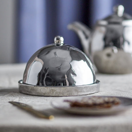 Shine Stoneware Butter Dish in silver with a mirror-bright domed lid, round knob handle, and hammered-texture base, shown on a table beside a teapot and side plate.