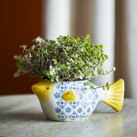 Wrenna Fish Stoneware Plant Pot in white and yellow with blue floral pattern, fin and fanned tail, holding a leafy green plant on a marble surface.