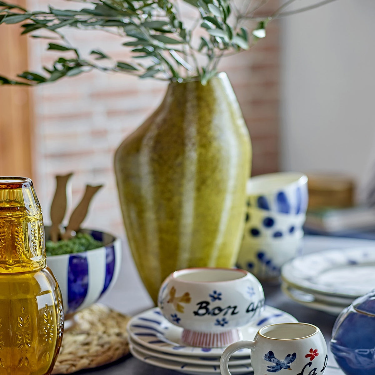 Abella Speckled Stoneware Vase in yellow, 35.5cm tall with an asymmetrical shoulder and vertical speckled bands, holding olive branches on a table with patterned ceramic bowls and cups.