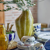 Abella Speckled Stoneware Vase in yellow, 35.5cm tall with an asymmetrical shoulder and vertical speckled bands, holding olive branches on a table with patterned ceramic bowls and cups.