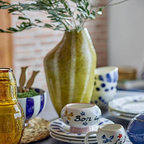 Abella Speckled Stoneware Vase in yellow, 35.5cm tall with an asymmetrical shoulder and vertical speckled bands, holding olive branches on a table with patterned ceramic bowls and cups.