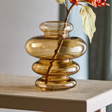 Kayn Glass Vase 19.5cm in Amber, with stacked rounded tiers in transparent glass, shown on a wooden table holding a leafy floral stem.