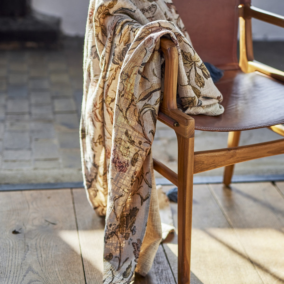 Yoti Floral Cotton Throw in multicoloured draped over a wooden armchair with tan leather seat, showing the all-over floral print and textured cotton weave.