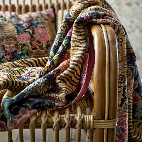 Lester Floral Cotton Throw in multicoloured floral and tiger-stripe print, draped over a rattan chair with a plain reverse side and matching patterned cushion visible beneath.