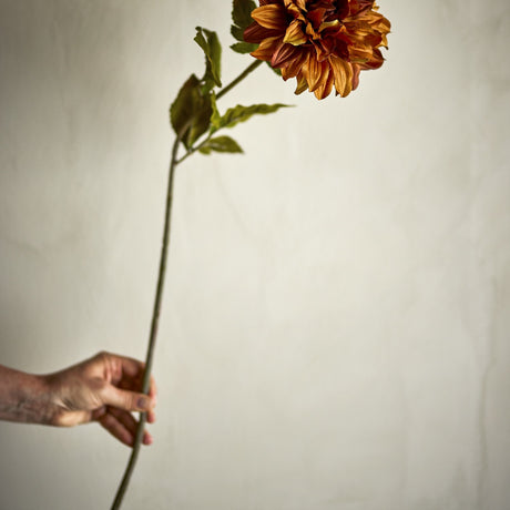 Chrysanthemum Artificial Stem 73cm in orange, held by hand against a plain light background, showing a layered bloom with leaves along a slender stem.