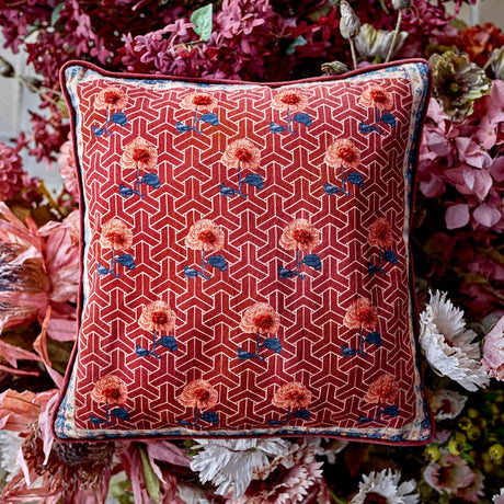 Zharai Geometric Cotton Cushion in red, square with piped edge, featuring a lattice pattern and floral rows, bordered in cream and blue, displayed among dried flowers.