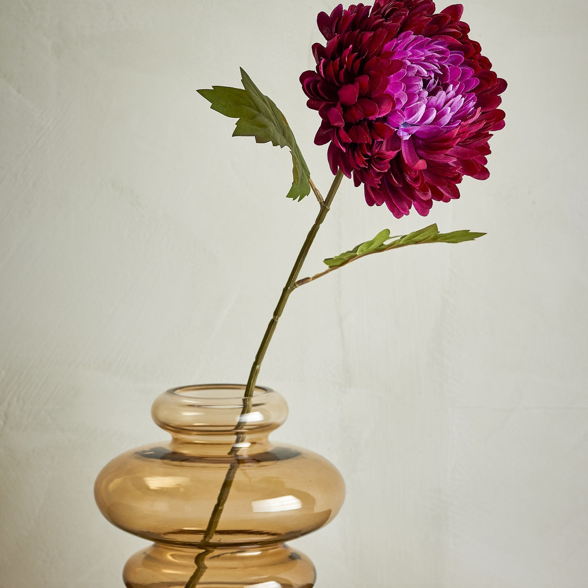 Chrysanthemum Artificial Stem in red, a single faux bloom with layered petals and green leaves, shown in an amber glass vase against a plain light background.