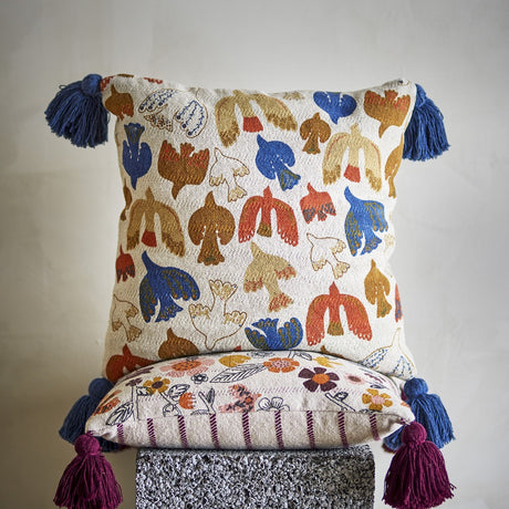 Anniversary Cotton Cushion in multi-coloured bird print with blue corner tassels, shown stacked on a floral striped cushion with burgundy tassels, plain background.