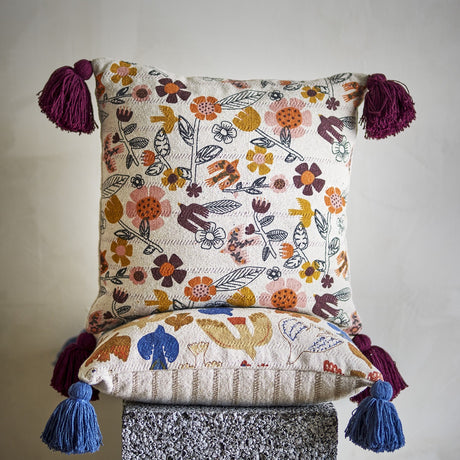 Vivielle Floral Cotton Cushion in multicoloured fabric with flowers, leaves and bird motifs, finished with burgundy and blue corner tassels, shown stacked on a textured stool.