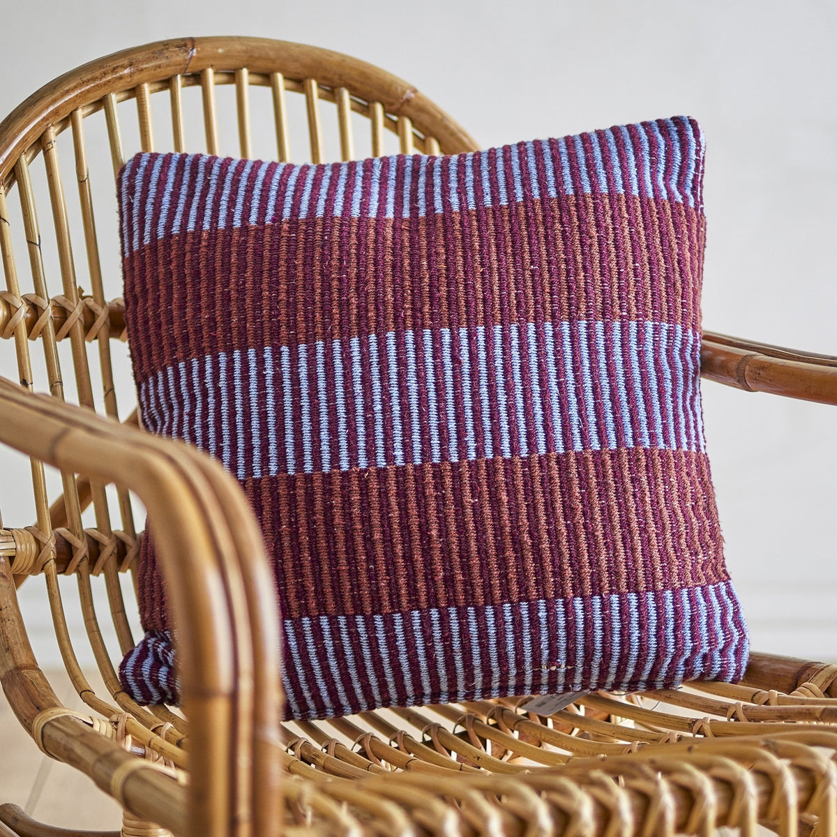 Isnel Brown Striped Cushion in multi tones, woven fabric with wide ribbed bands and fine pinstripes, shown on a rattan chair.
