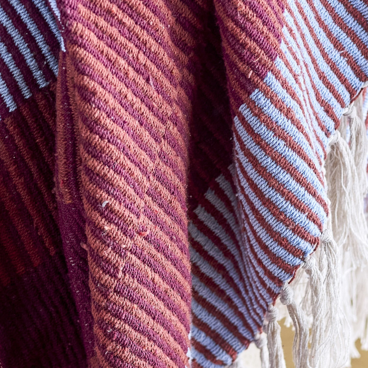 Close-up of the Isnel Red Striped Throw, a woven fabric throw with diagonal pinstripes in maroon, terracotta, and blue, showing ribbed texture, folds, and knotted yarn fringe.