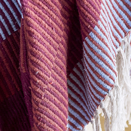 Close-up of the Isnel Red Striped Throw, a woven fabric throw with diagonal pinstripes in maroon, terracotta, and blue, showing ribbed texture, folds, and knotted yarn fringe.
