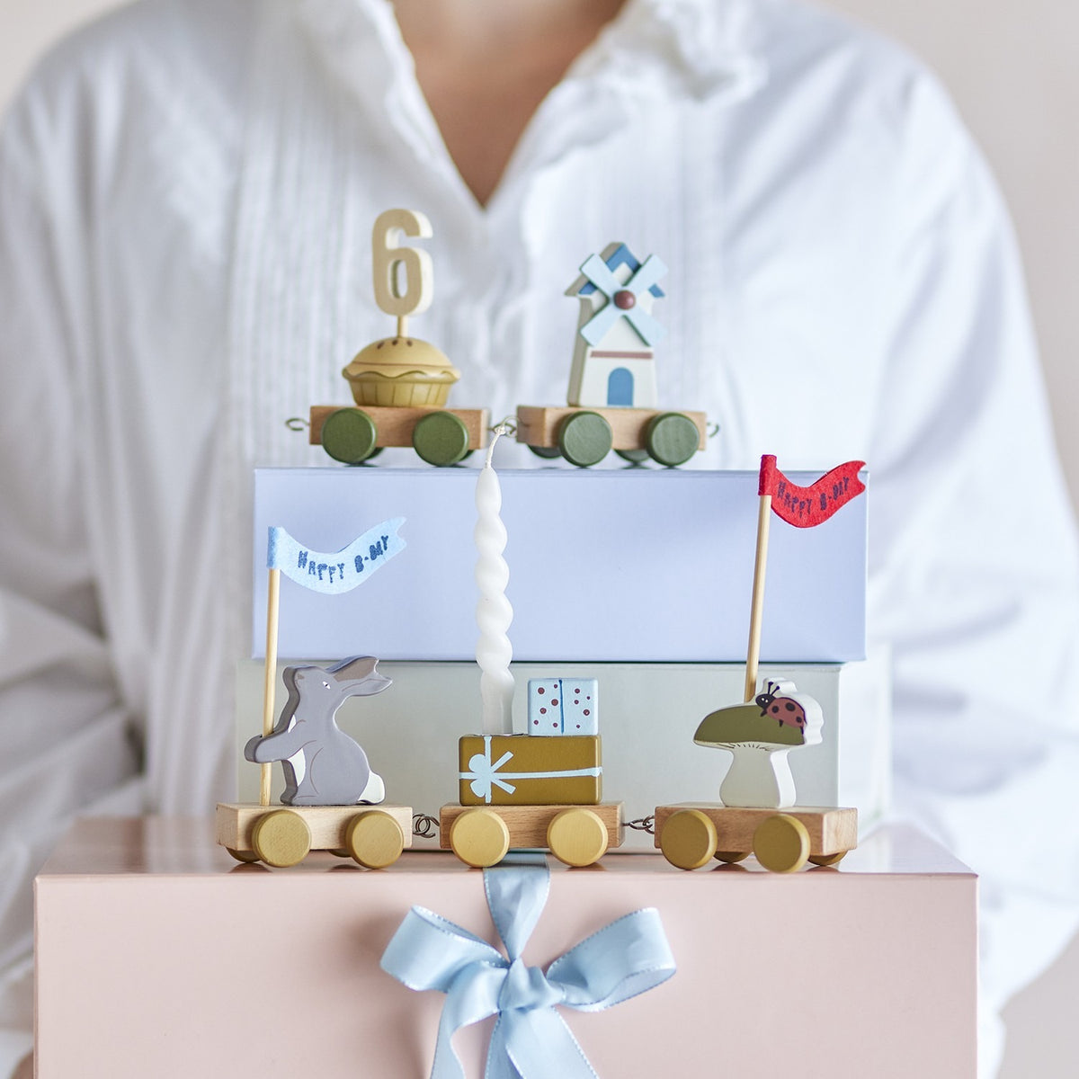 Alfi Birthday Decoration wooden train with rabbit, gift, windmill and mushroom wagons, number "6" topper on cake, held atop pink gift box with blue ribbon.