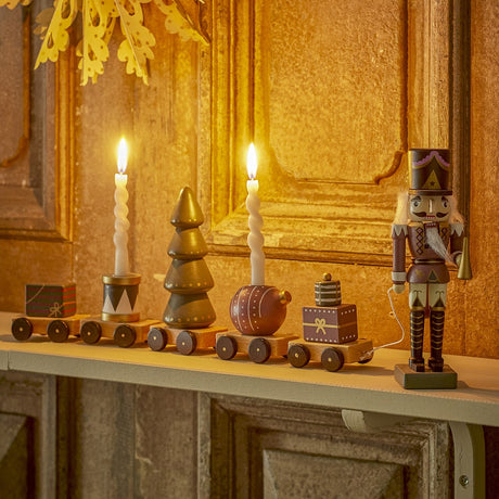 Vadim Christmas Decoration 18.5cm: a wooden nutcracker figure pulling five wagons with parcels, a drum, tiered tree and bauble, displayed on a mantel with lit candles.