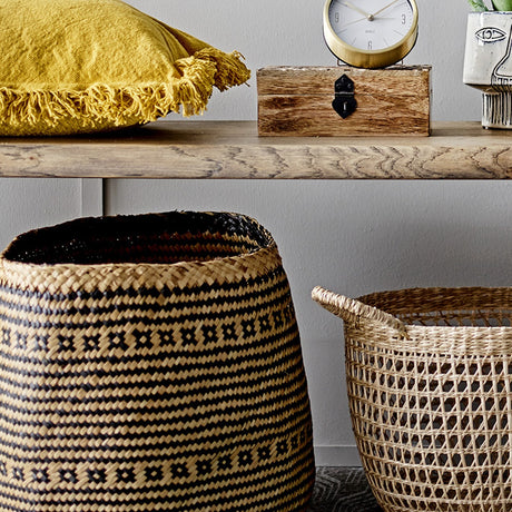 Heddy Seagrass Baskets Set of 2 in Black, woven storage baskets with zigzag and cross motif patterns, shown under a wooden shelf in a living room setting.