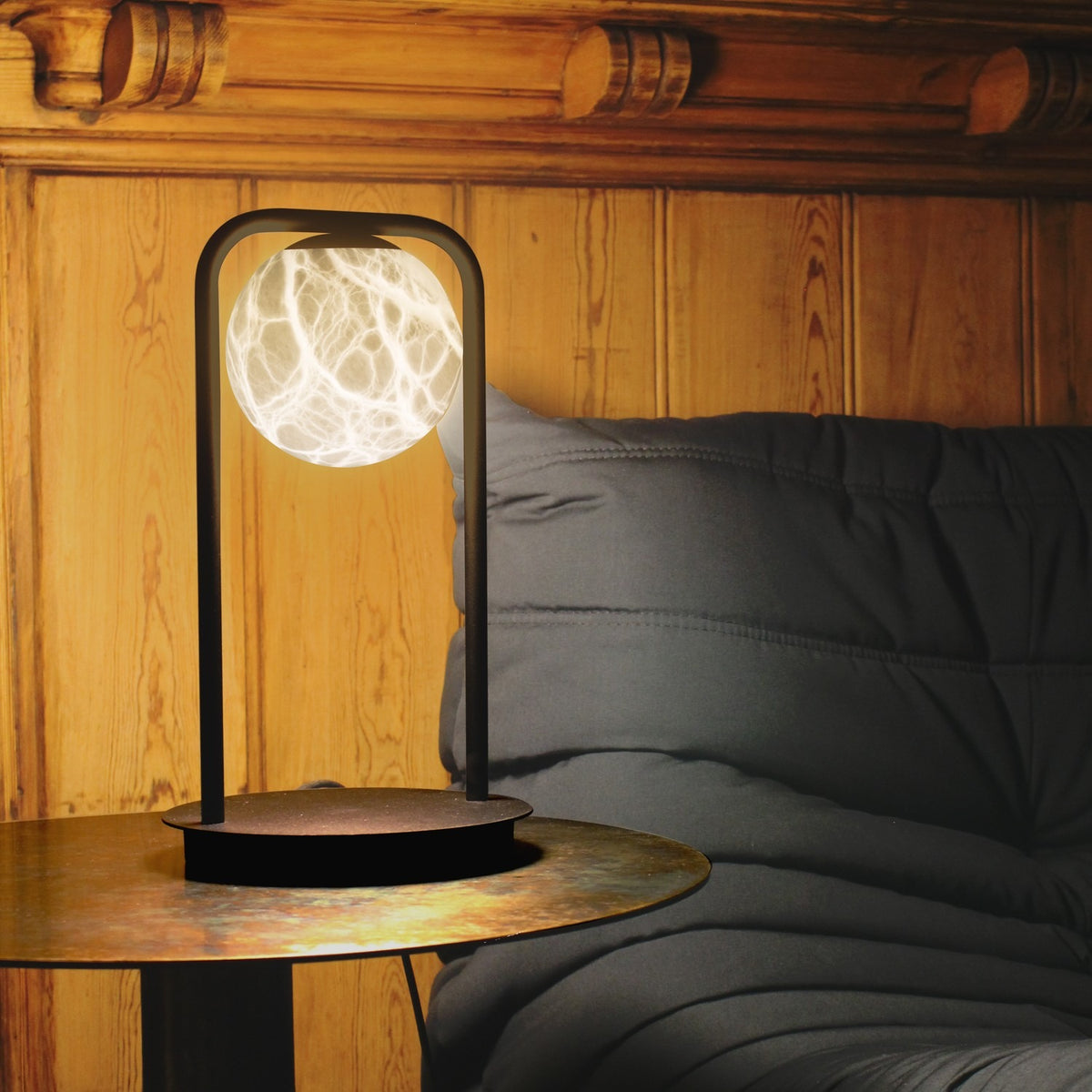 Halvessa LED Alabaster Table Lamp in black, with a glowing veined alabaster globe on a slim arched iron frame, shown on a table beside a grey sofa in a wood-panelled room.