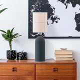 Rania table lamp with cylindrical anthracite concrete base and white cream fabric shade with stitched grid lines, styled on a wooden sideboard with books and a plant.