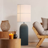 Rania Floor Lamp with a tall rounded anthracite concrete base and white cream cylindrical fabric shade, shown lit beside a wooden sofa in a living room.