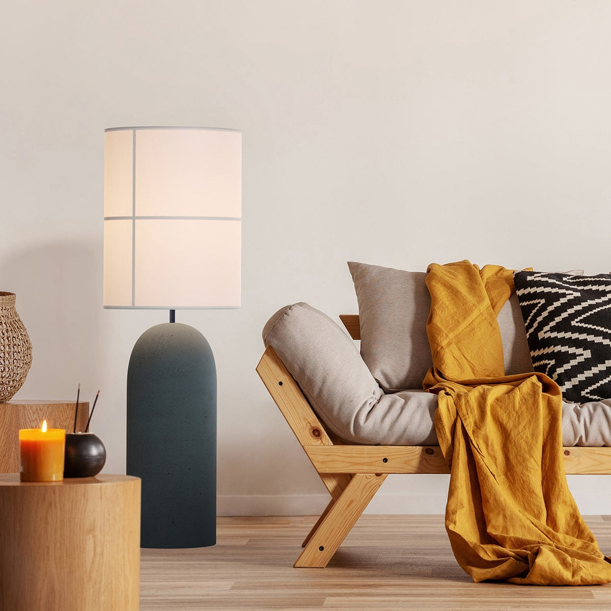 Rania Floor Lamp with a tall anthracite concrete column base and white cream fabric cylindrical shade, shown beside a wooden sofa in a living room setting.