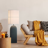 Rania Floor Lamp with a tall anthracite concrete column base and white cream fabric cylindrical shade, shown beside a wooden sofa in a living room setting.