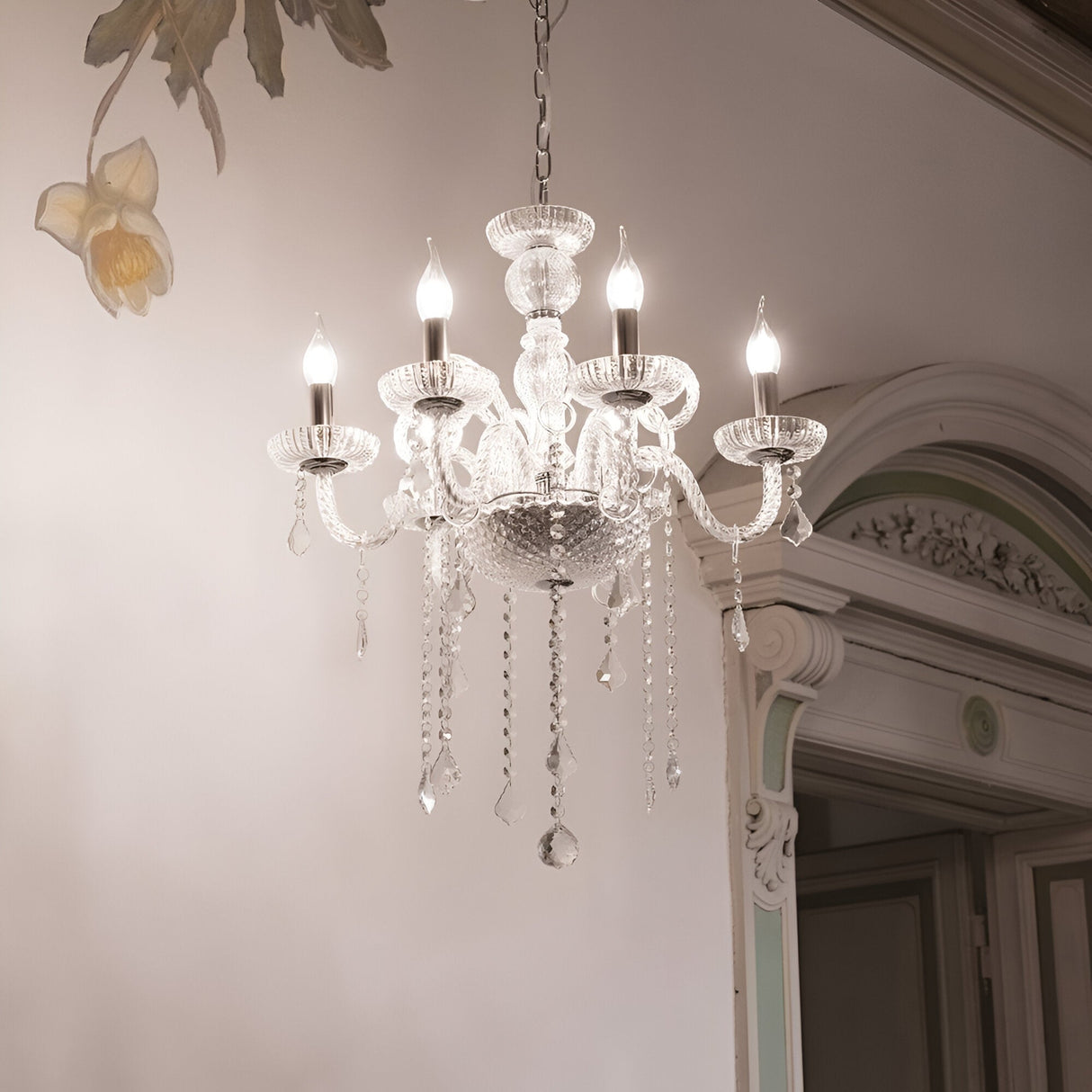 The Etherlight Crystal 6 Light Chandelier - Chrome gracefully hangs from the ceiling, highlighted by its illuminated bulbs, detailed glasswork, and elegant dangling ornaments. It complements a room adorned with ornate molding and floral wall designs, merging classic elegance with modern design elements.